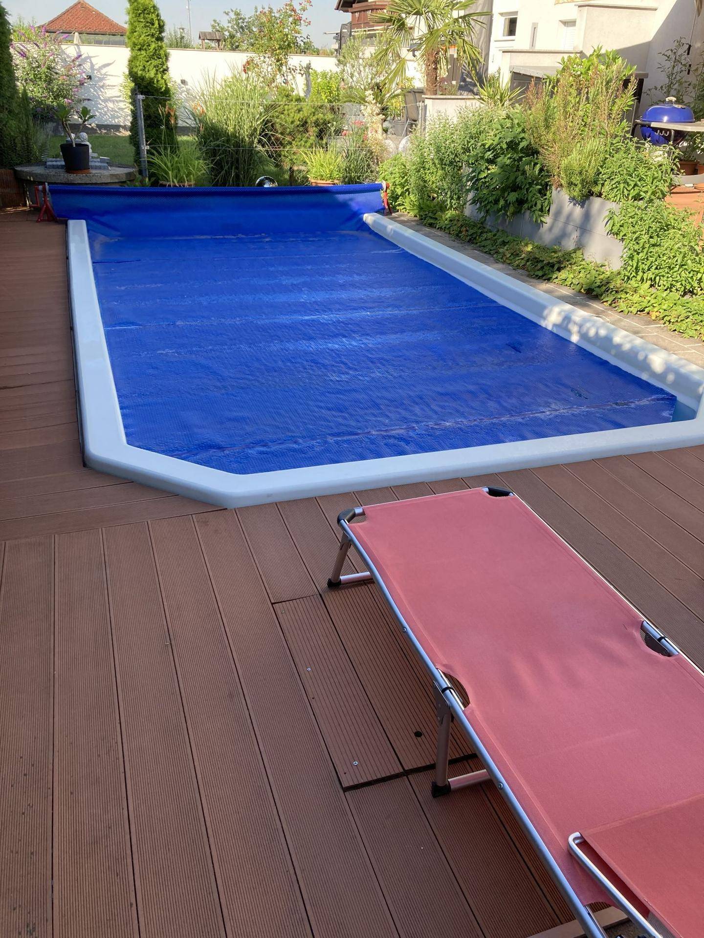 Außenpool mit blauer Abdeckung, umgeben von einer Holzterrasse und Garten.