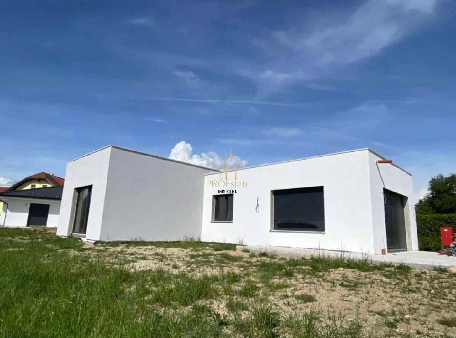 Moderne, weiß verputzte Neubaufassade mit Flachdach und großen Fenstern unter blauem Himmel.