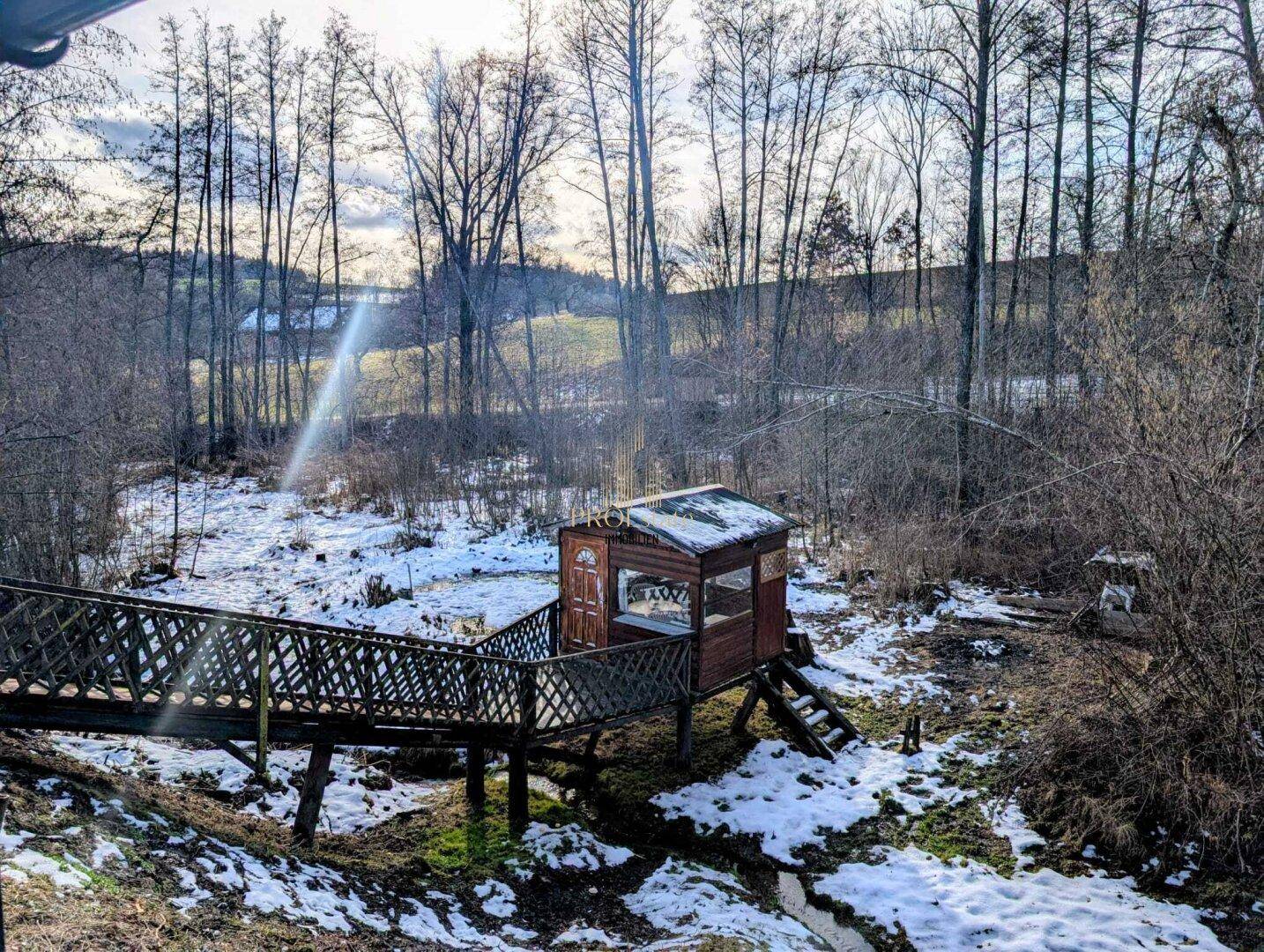 Winterliche Landschaft mit einem kleinen Holzhaus und einer Brücke, umgeben von Bäumen und Schnee.