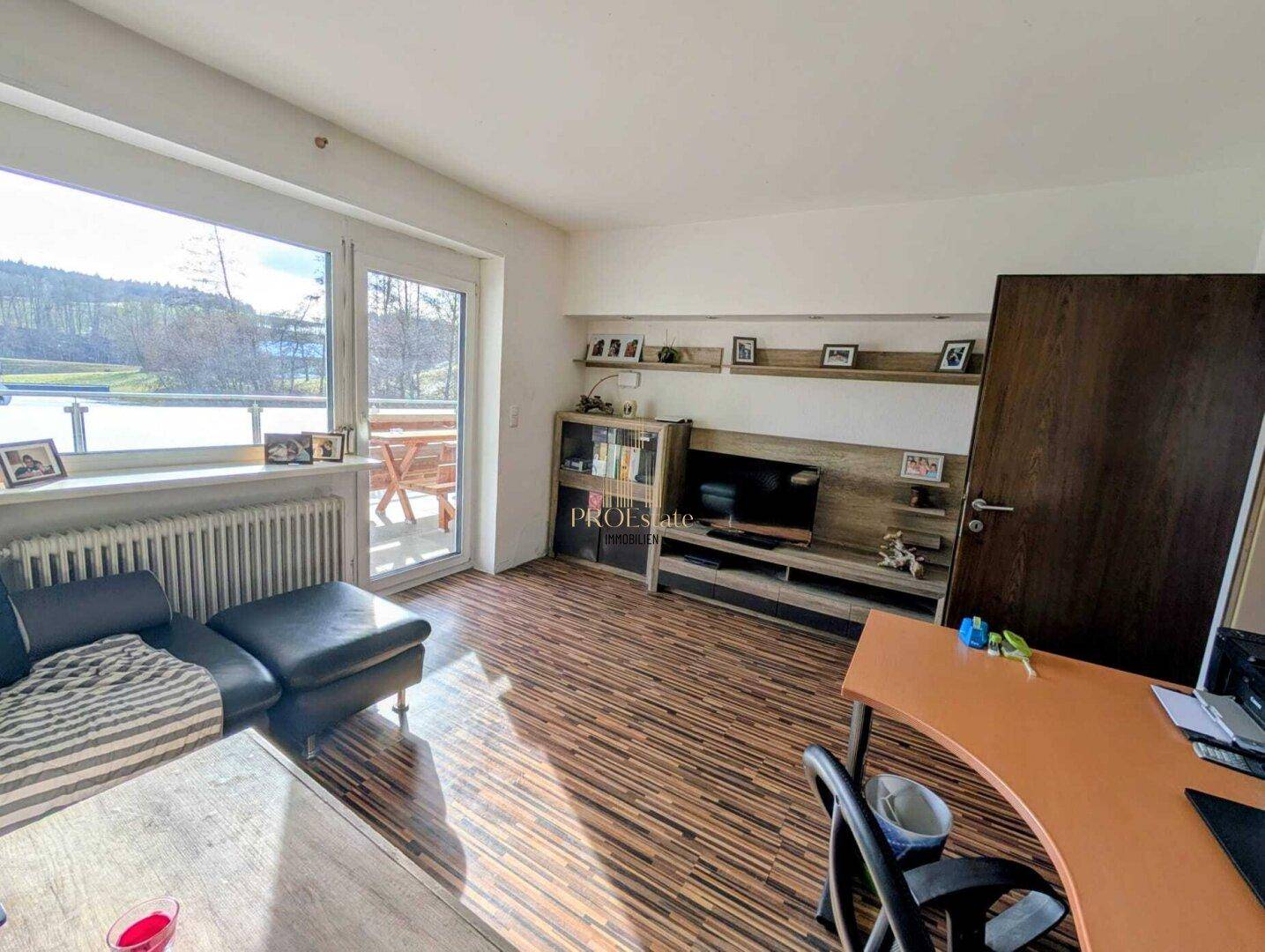 Helles Wohnzimmer mit Holzboden, großem Fenster und Zugang zu einem Balkon.