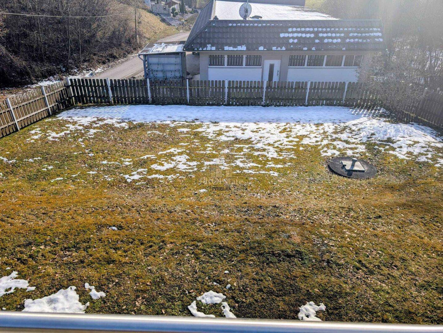Gartenbereich mit Rasenfläche und leichtem Schneefall, umzäunt und mit Blick auf die Umgebung.