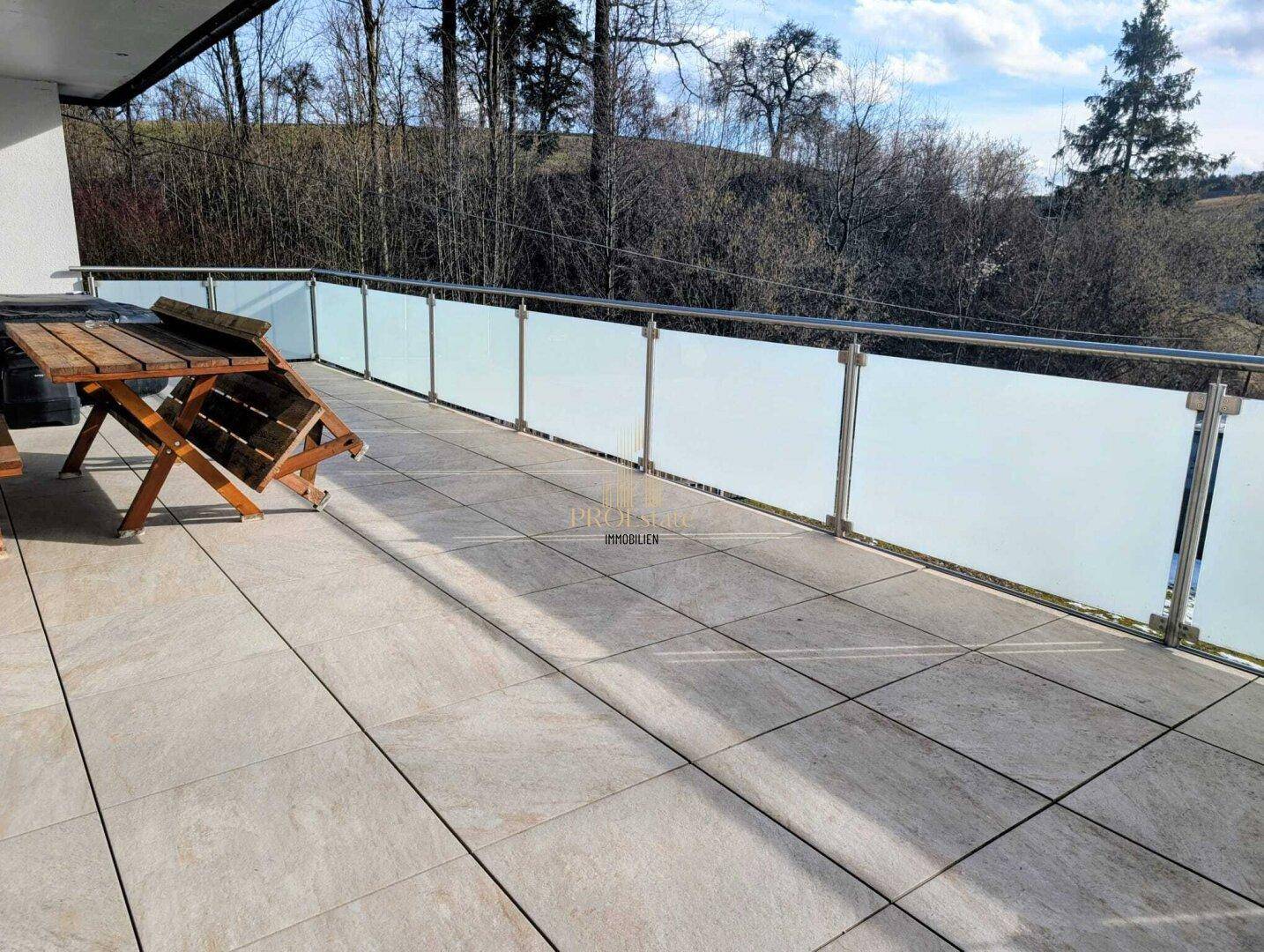 Große geflieste Terrasse mit Glasgeländer und Holztisch, umgeben von Bäumen.