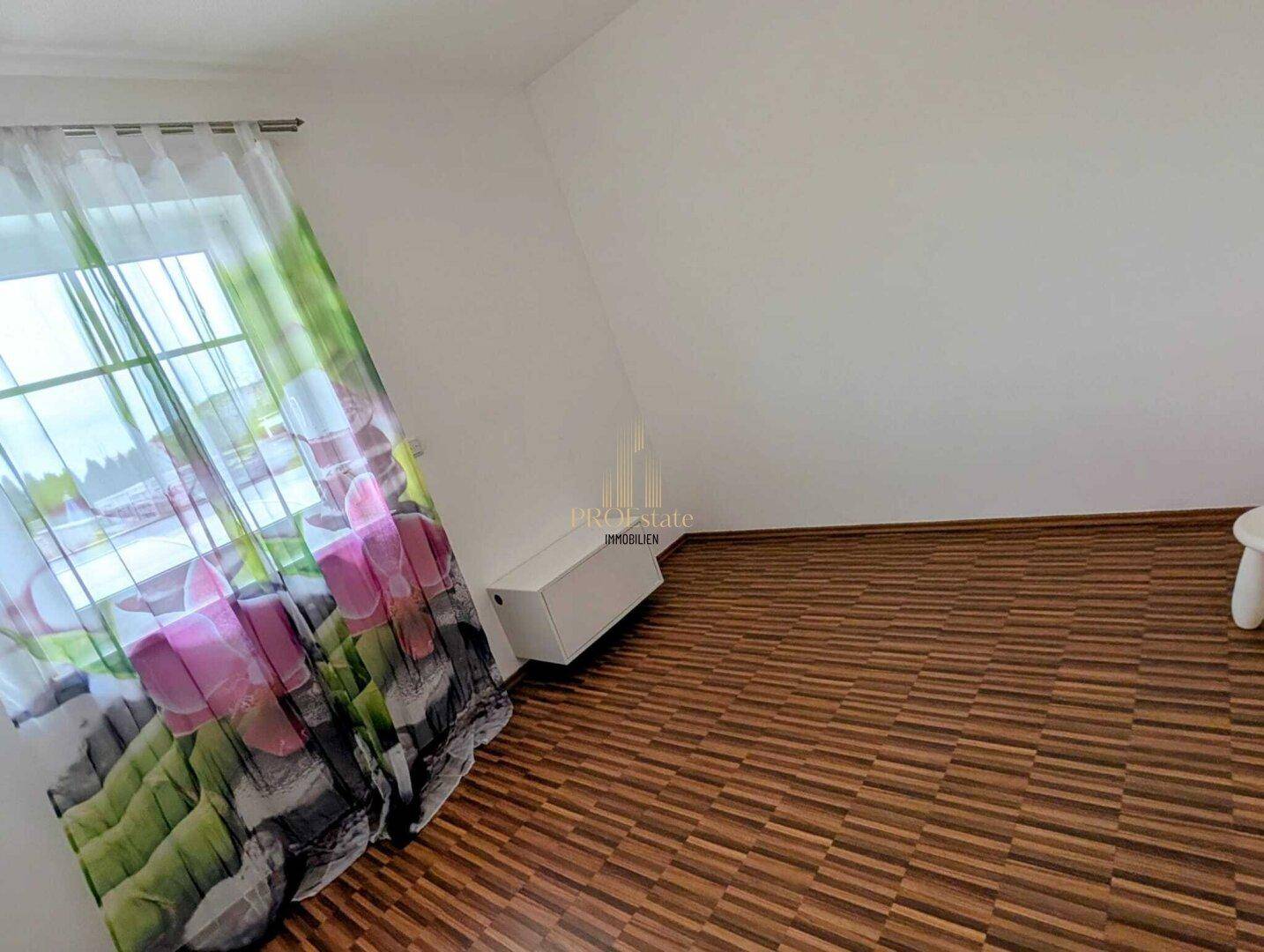 Helles Schlafzimmer mit Laminatboden und einem Fenster mit floralem Vorhang.