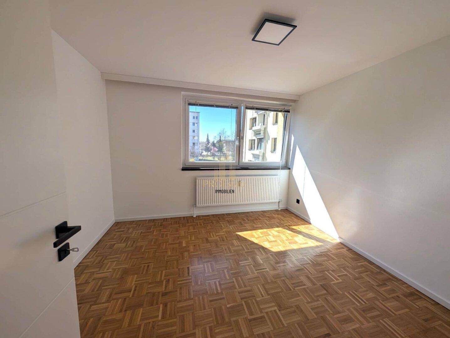 Leeres Zimmer mit Parkettboden und großem Fenster, das viel Tageslicht hereinlässt und eine angenehme Atmosphäre schafft.