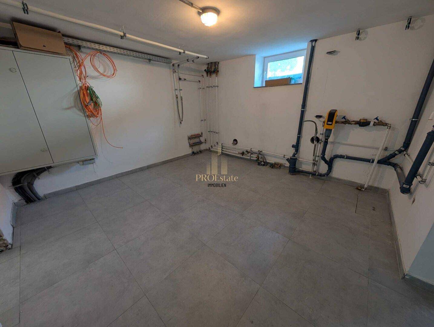 Technikraum im Keller mit Installationen, Fliesenboden und kleinem Fenster.