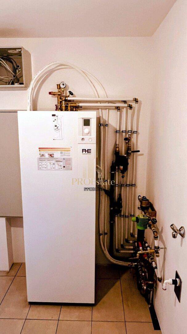 Hauswirtschaftsraum mit moderner Wärmepumpe und sichtbaren Rohrleitungen auf Fliesenboden.