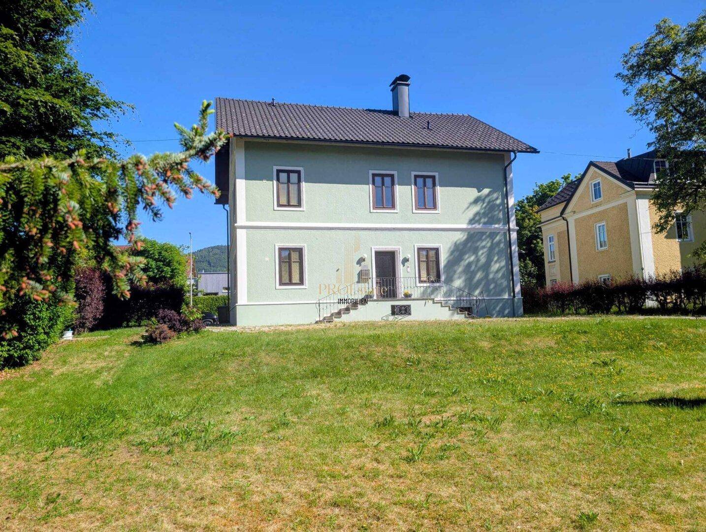Seitenansicht des Hauses mit weitläufigem Rasen und umgebender Natur unter blauem Himmel.