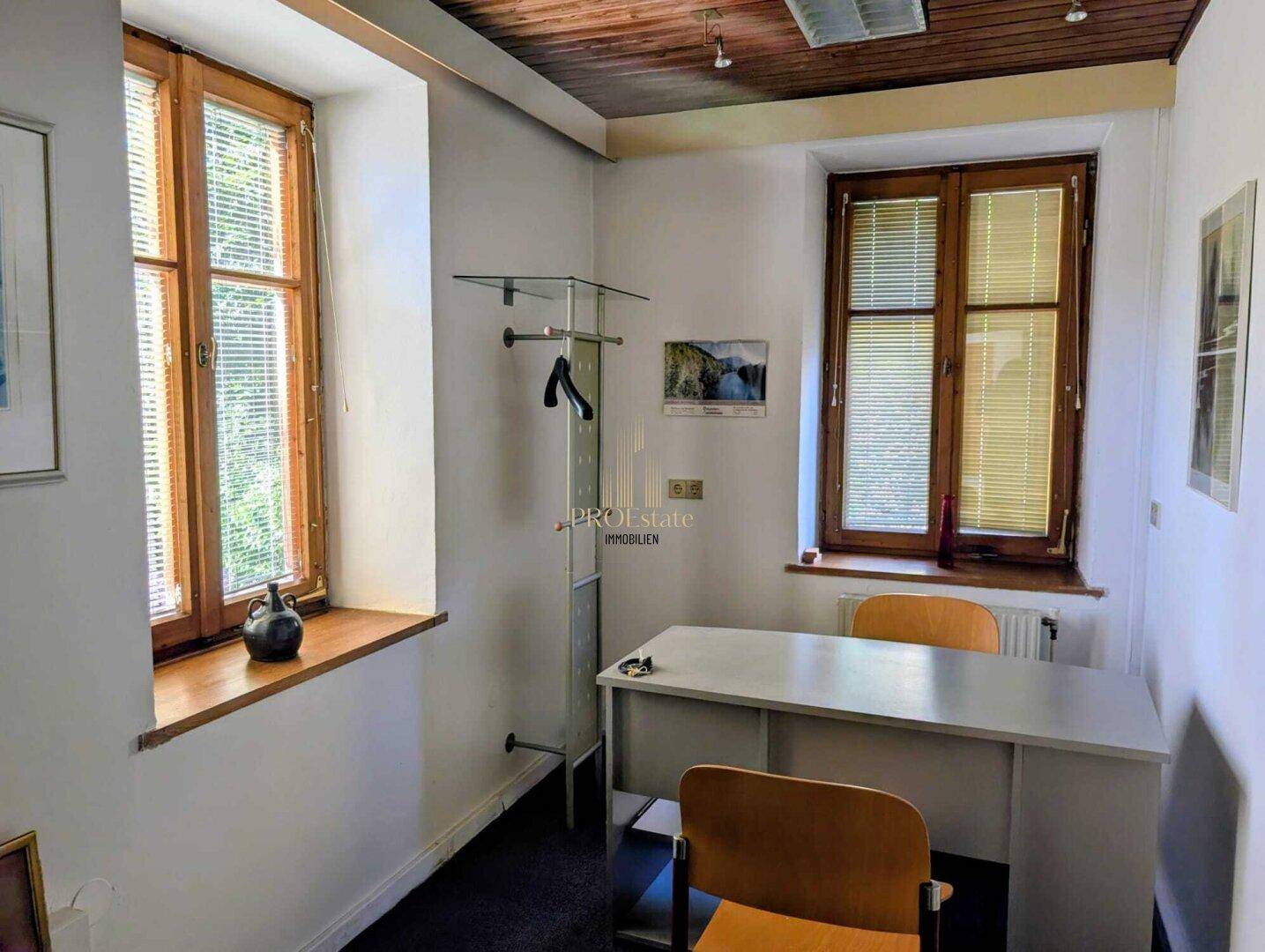Kleines Büro mit zwei Fenstern, Schreibtisch und Garderobe, ausgestattet mit Holzdecke.