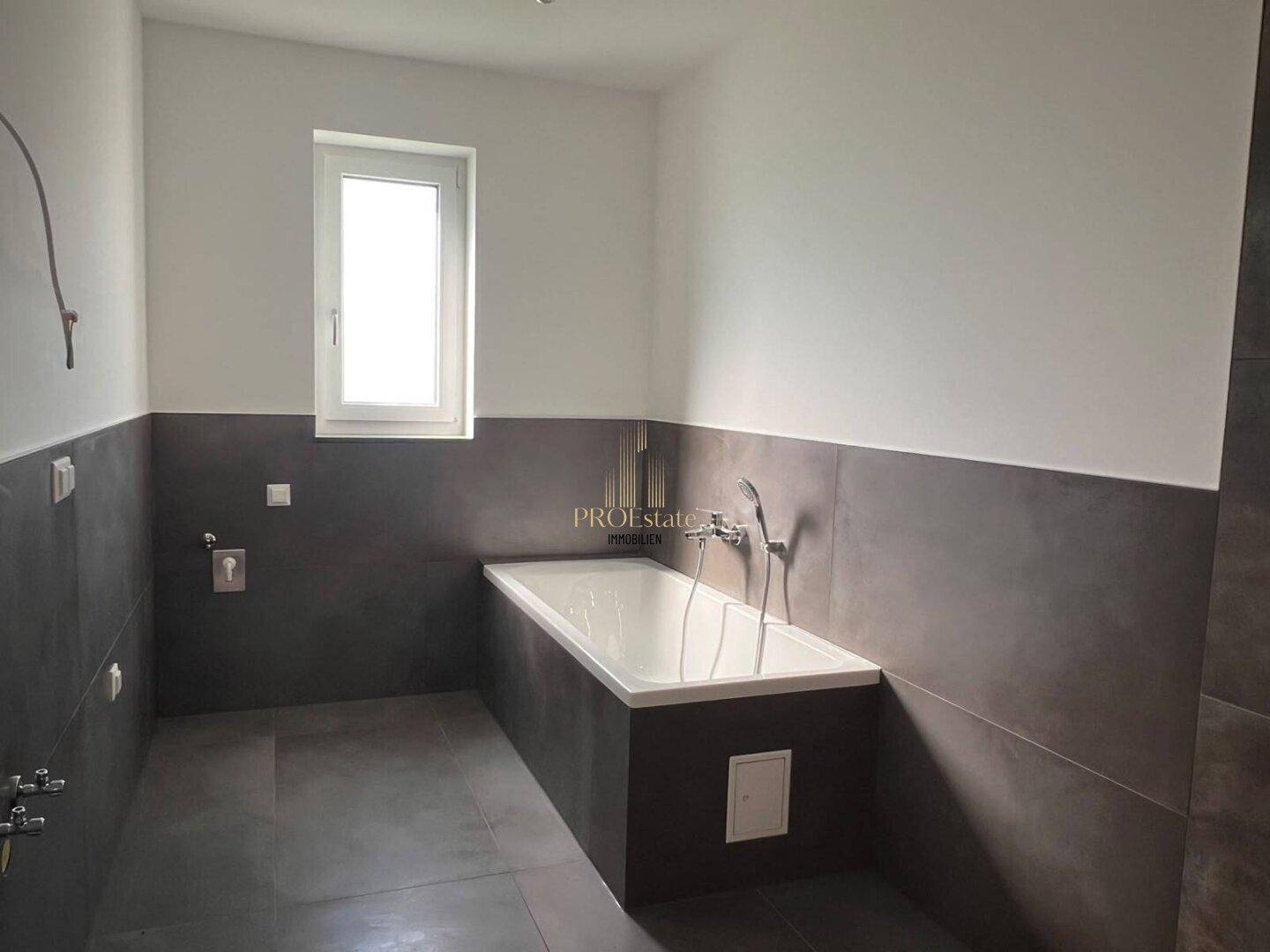 Modernes Badezimmer mit Badewanne, dunklen Fliesen und Fenster für natürliches Licht.