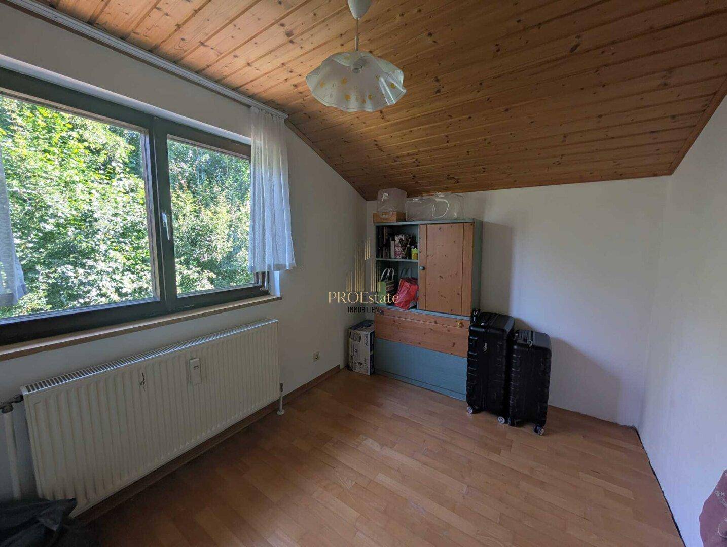 Dachzimmer mit Holzdecke, Holzboden und einem Fenster mit Blick ins Grüne.