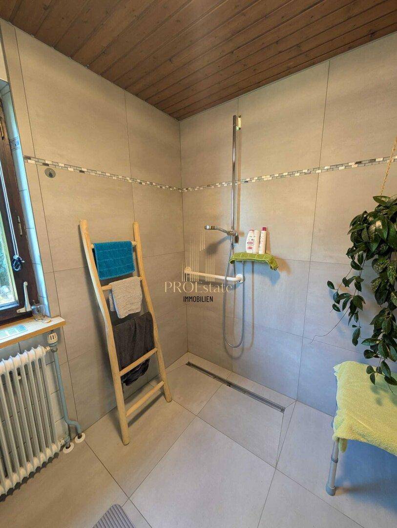 Gefliestes Badezimmer mit ebenerdiger Dusche und Holzdecke, hell durch das Fenster.