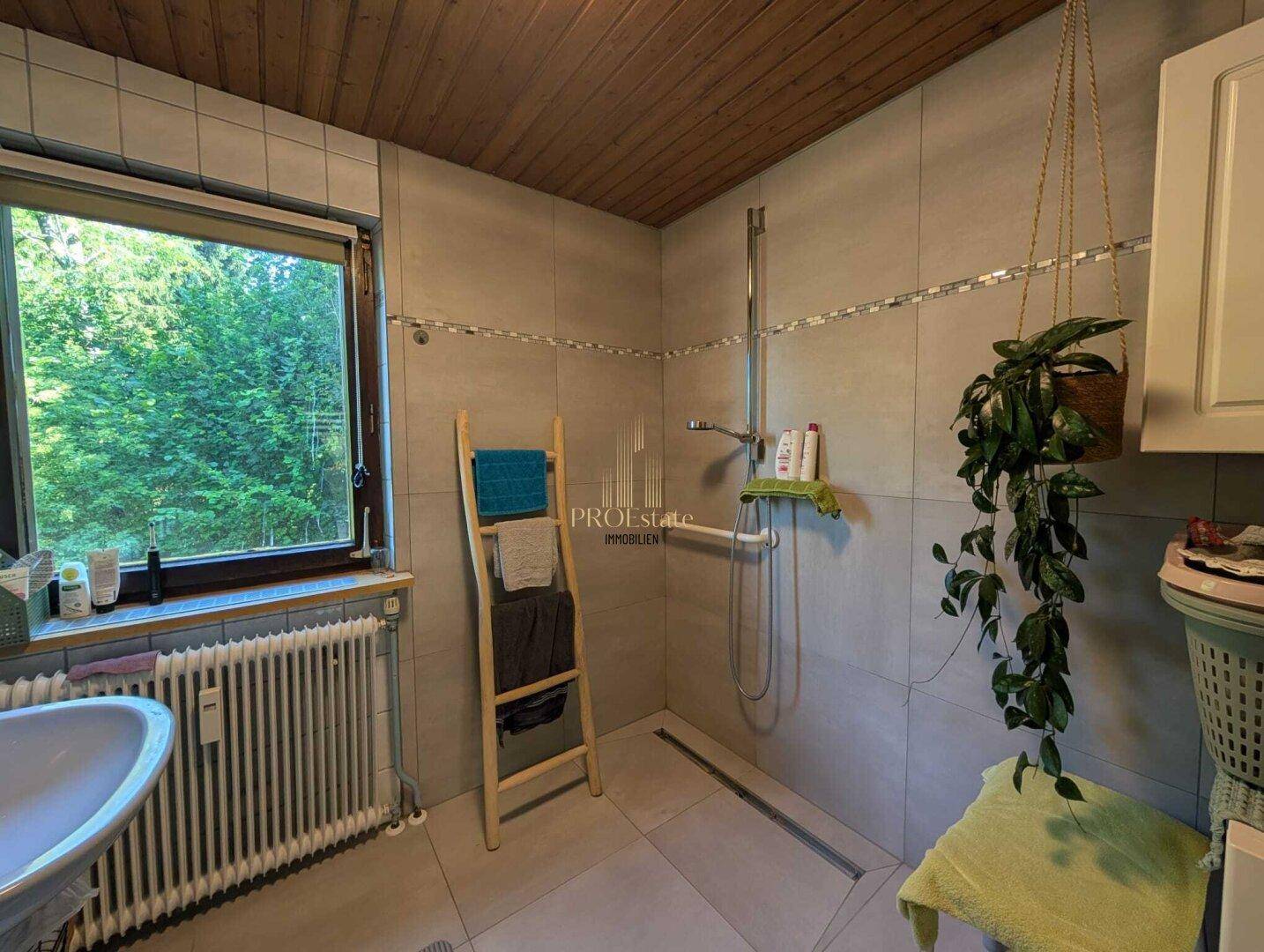 Badezimmer mit gefliester Dusche, Holzdecke und einem Fenster mit Blick ins Grüne.