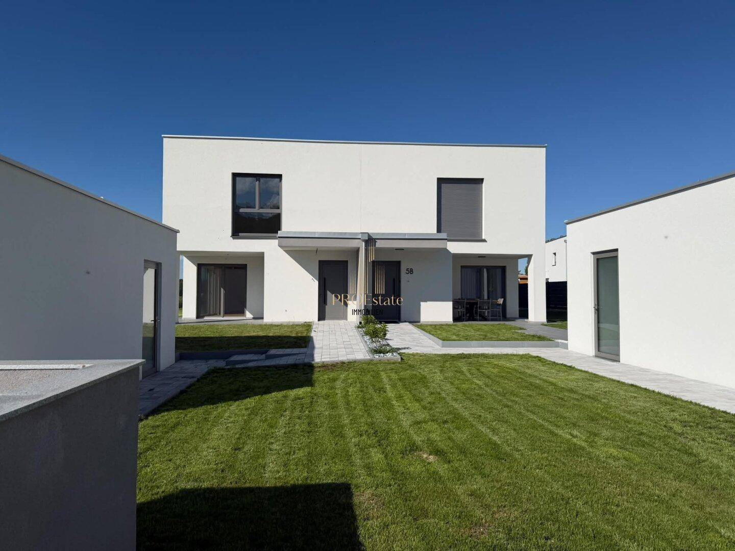 Moderne weiße Doppelhaushälfte mit klaren Linien und gepflegtem Vorgarten unter blauem Himmel.