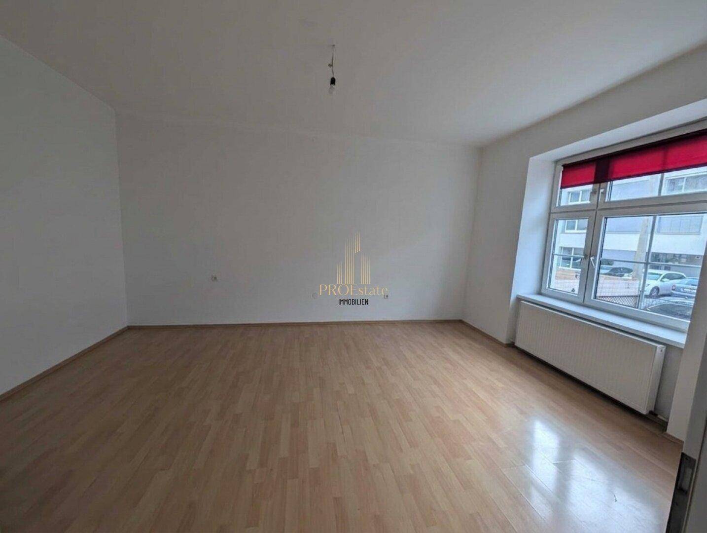 Geräumiges Wohnzimmer mit Parkettboden und einem großen Fenster mit rotem Rollo.