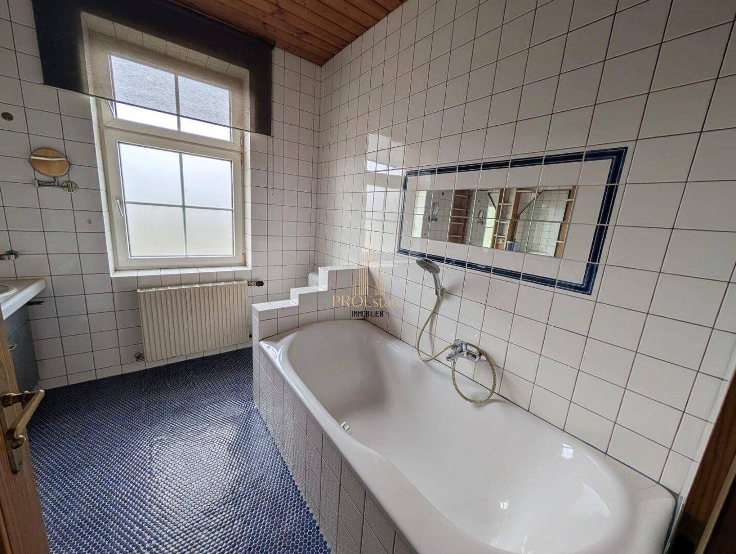 Badezimmer mit eingebauter Badewanne, weißen Wandfliesen und blauen Bodenfliesen sowie einem Fenster.