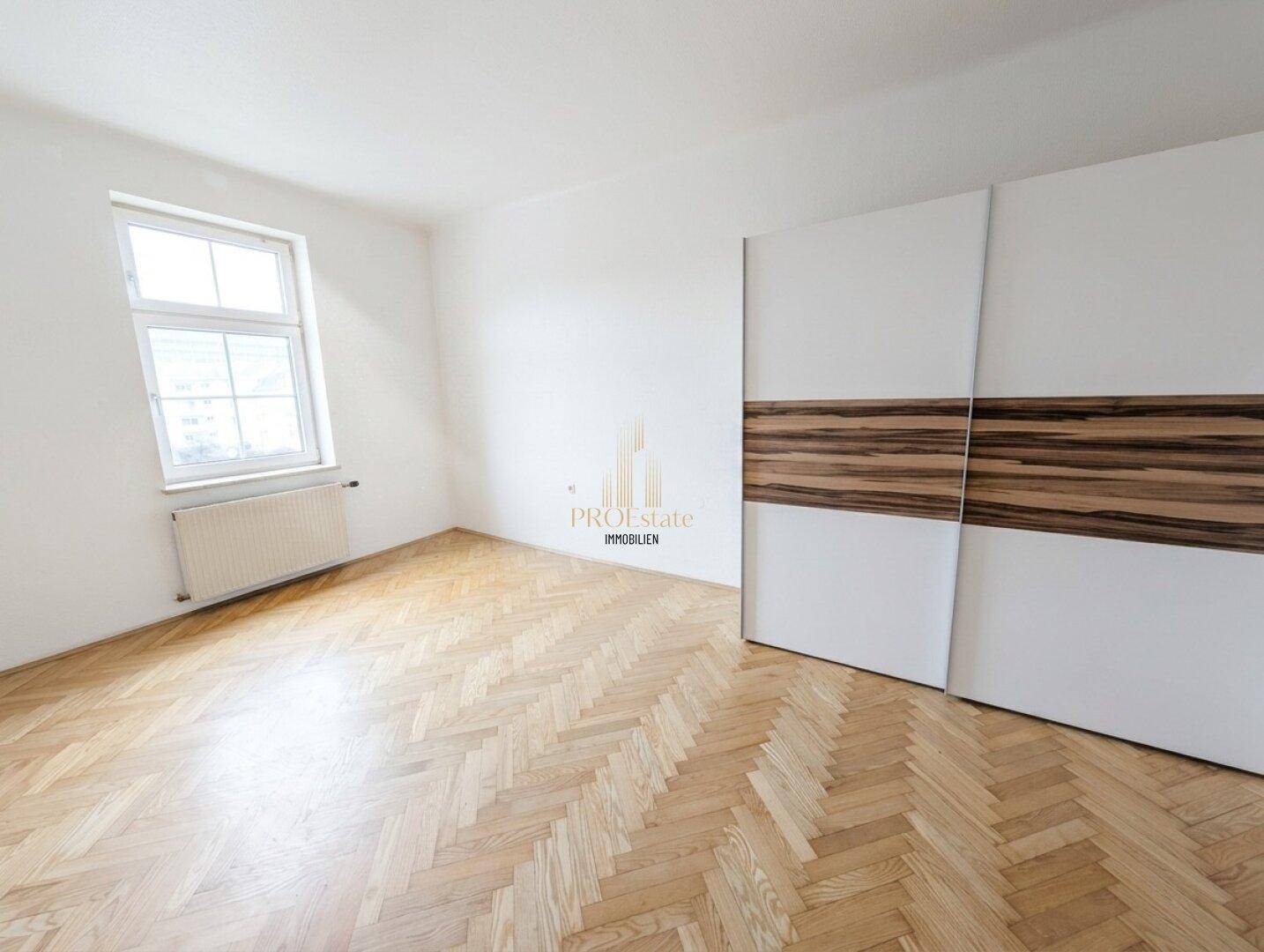Helles Schlafzimmer mit Fischgrätparkett, großem Fenster und integriertem Kleiderschrank in modernem Design.