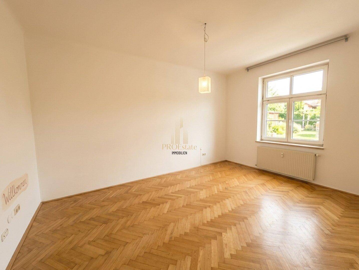 Geräumiges Zimmer mit Fischgrätparkett, hellen Wänden und einem Fenster mit Blick ins Grüne.