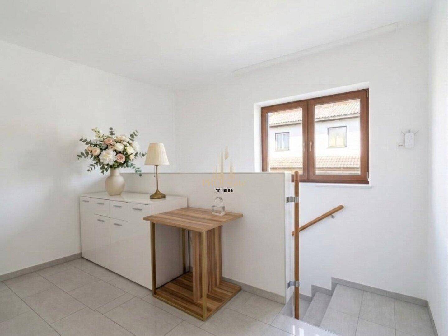Heller Treppenabsatz mit Fenster, Kommode und dekorativen Blumen.