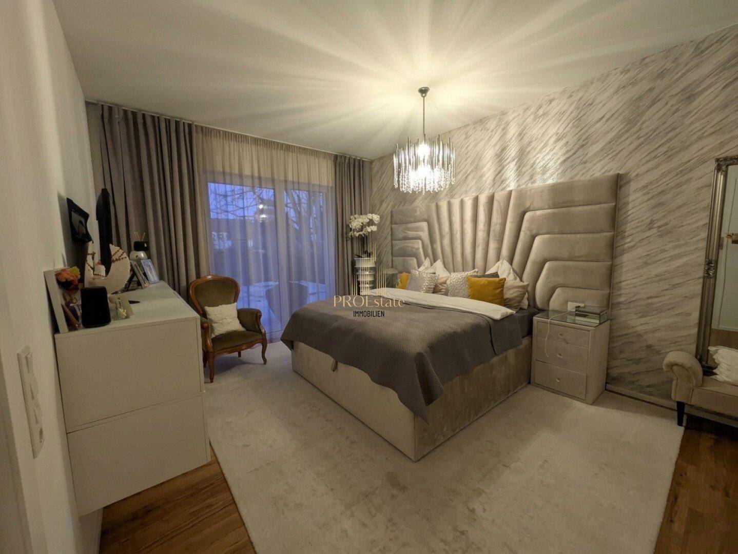 Elegantes Schlafzimmer mit großem gepolstertem Kopfteil, Kronleuchter und Zugang zum Außenbereich.