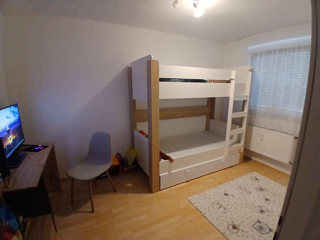 Kinderzimmer mit Etagenbett, Schreibtisch und Fenster mit Jalousien, auf hellem Holzboden.