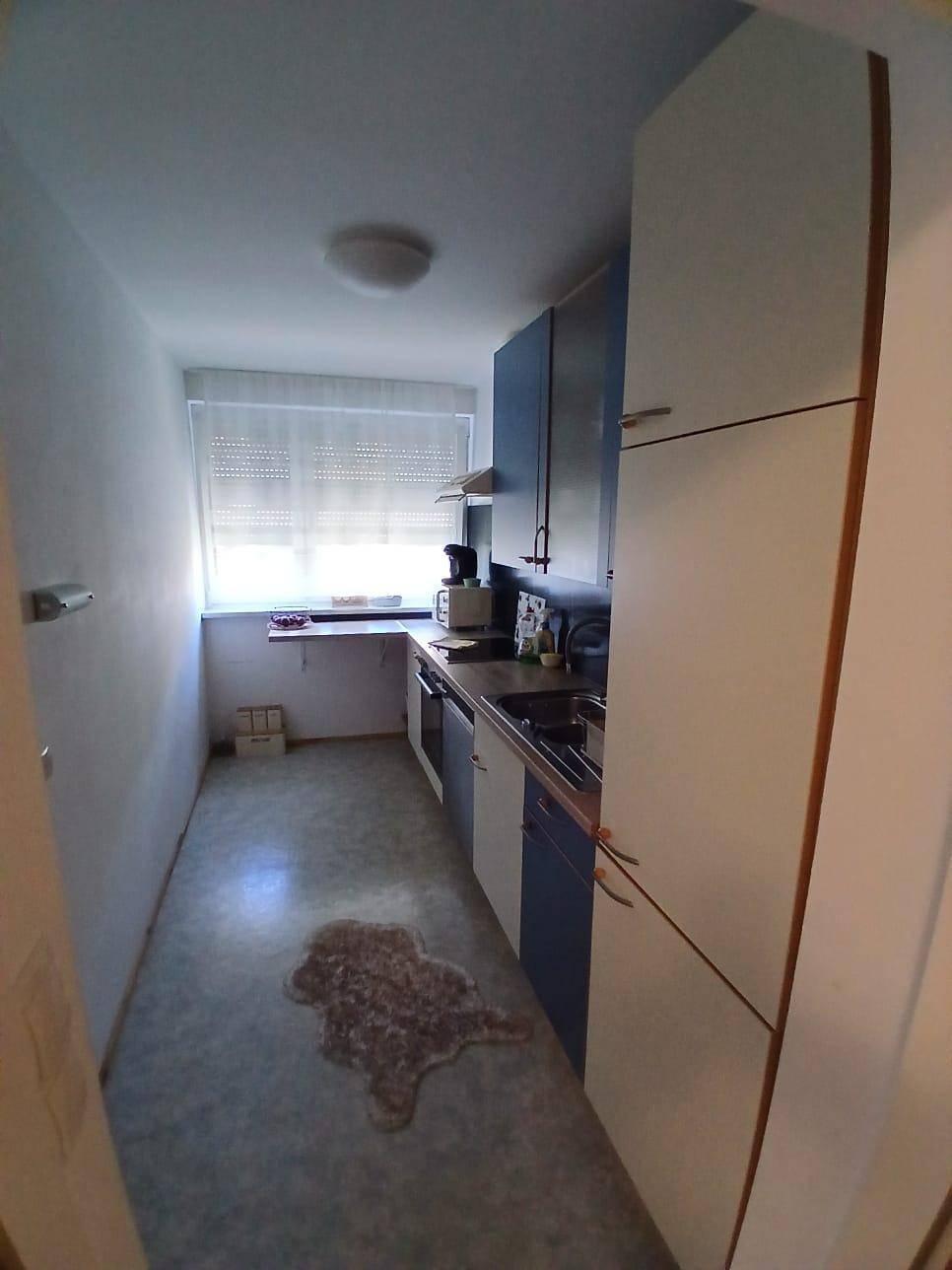 Schmale Küche mit blauen und weißen Schränken, Spüle, Fenster und integrierten Geräten.