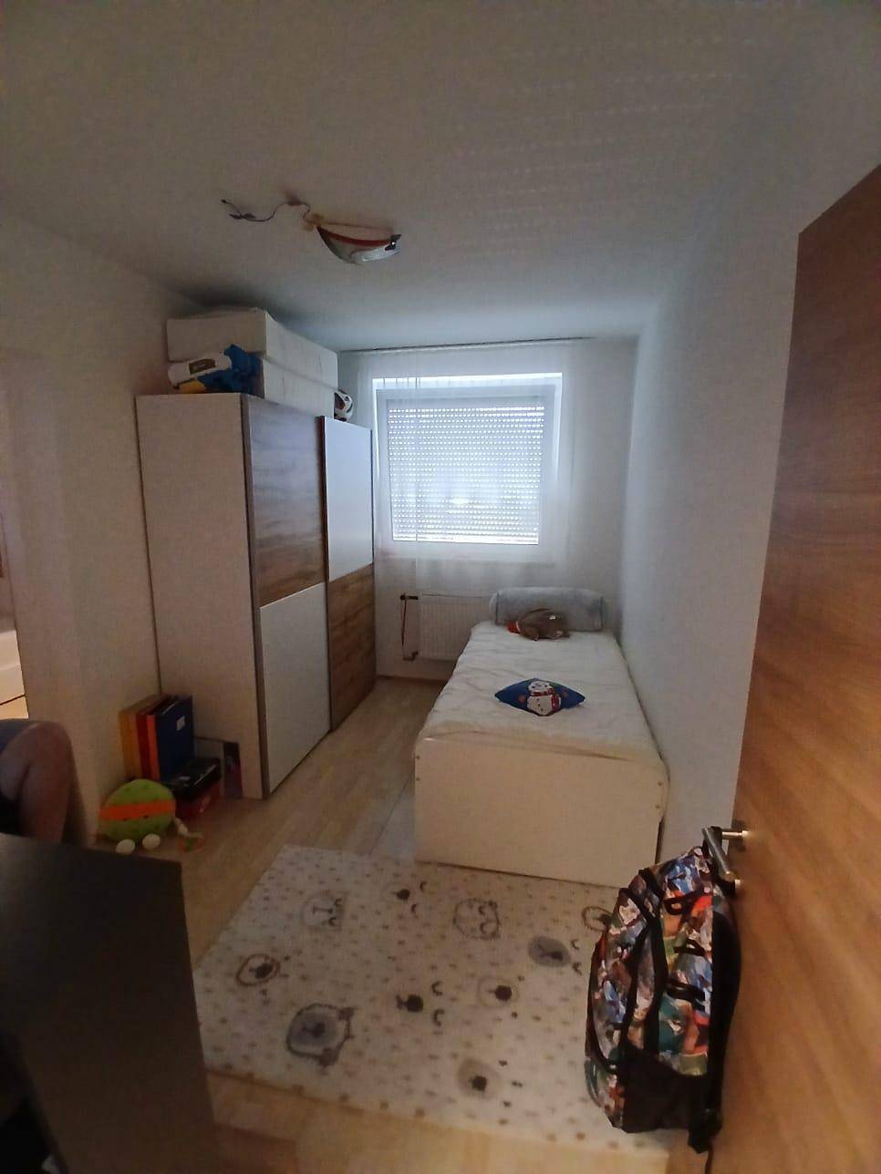 Kinderzimmer mit Einzelbett, Kleiderschrank und Fenster mit Jalousien, auf hellem Holzboden.
