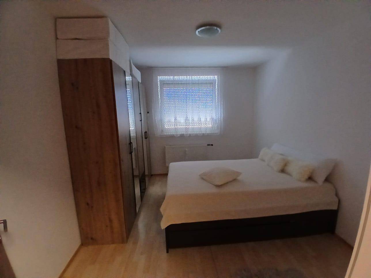 Schlafzimmer mit Doppelbett, großem Kleiderschrank und Fenster mit Jalousien, auf hellem Holzboden.