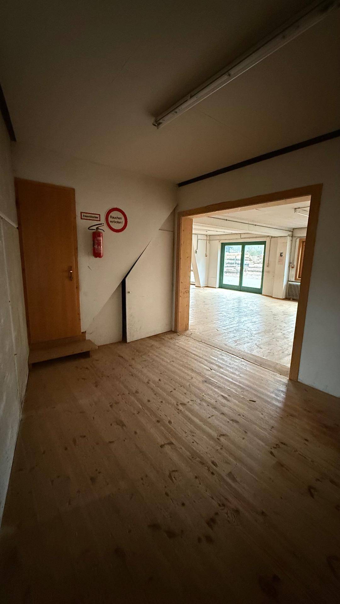Übergangsbereich mit Holzdielenboden und einer Holztür, führt in einen größeren Raum mit Tageslicht.