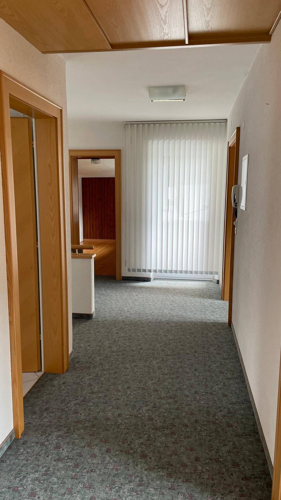 Langer Flur mit Teppichboden, Holzdecke und einem großen Fenster mit vertikalen Jalousien.