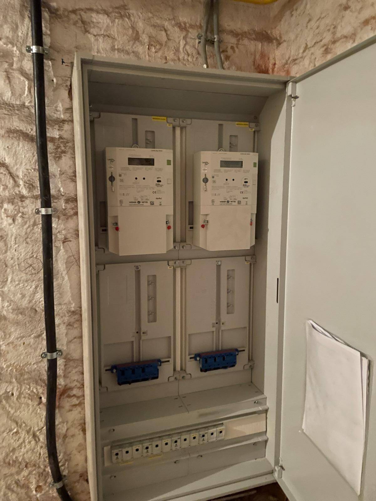 Offener Elektroschrank mit zwei Stromzählern vor einer unverputzten Ziegelwand im Kellerbereich.