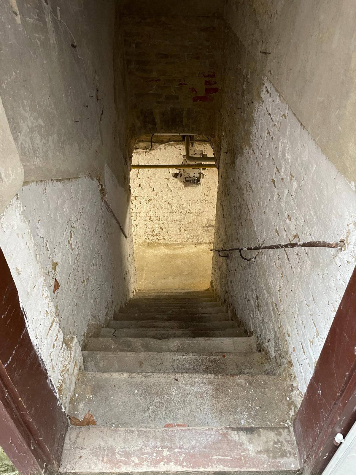 Betontreppe führt in einen dunklen Keller hinunter, umgeben von rauen, weiß gestrichenen Ziegelwänden.