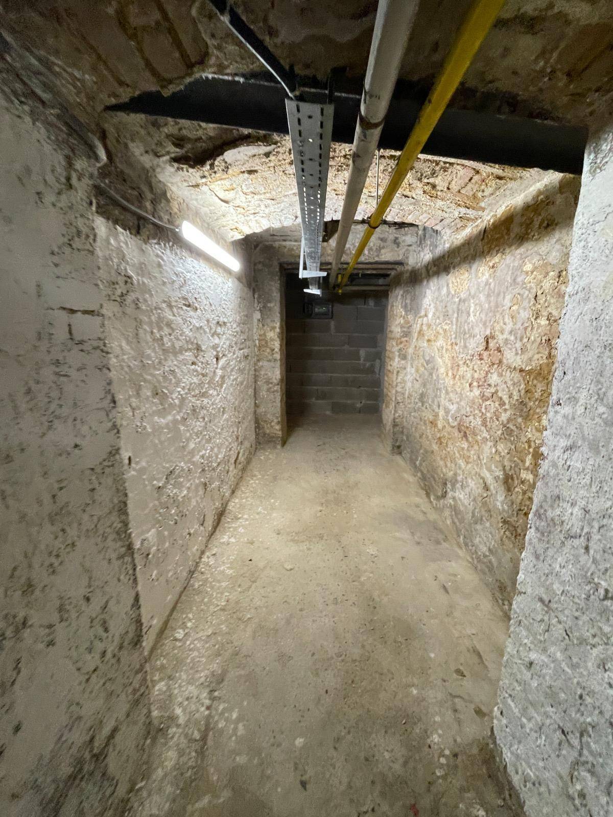 Langer, schmaler Kellergang mit rauen Steinwänden, Betonboden und einer Treppe am Ende.
