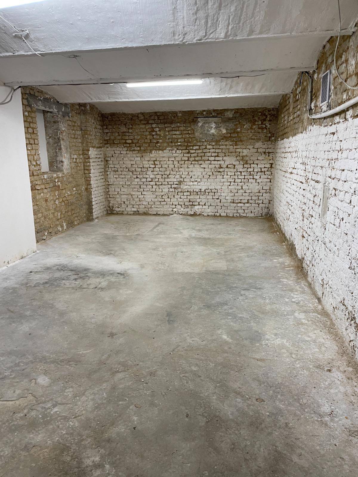 Leerer Kellerraum mit teilweise weiß gestrichenen Ziegelwänden, Betonboden und Deckenbeleuchtung.
