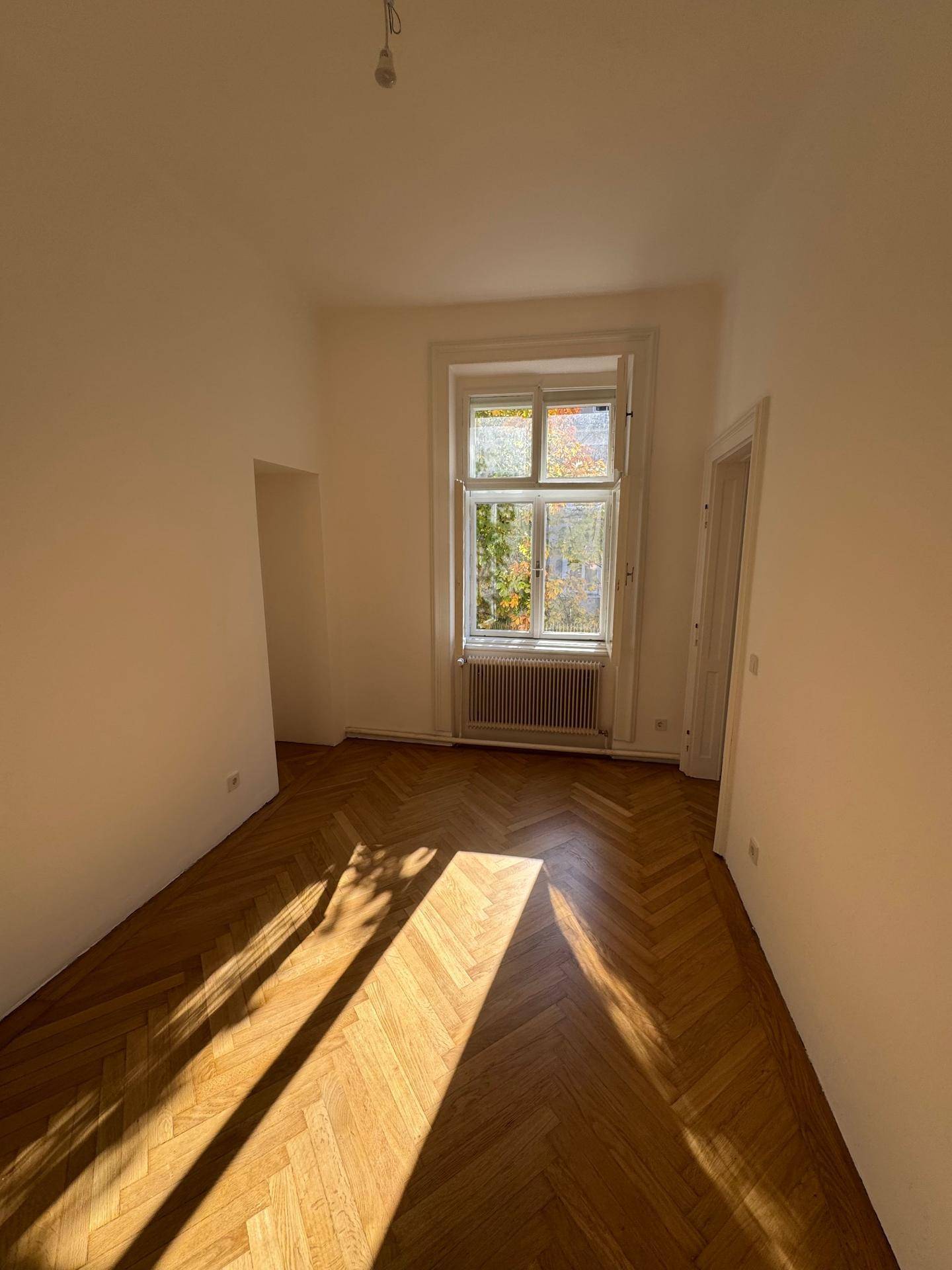 Lichtdurchfluteter Raum mit großen Fenstern, elegantem Fischgrätparkett und frisch gestrichenen weißen Wänden.