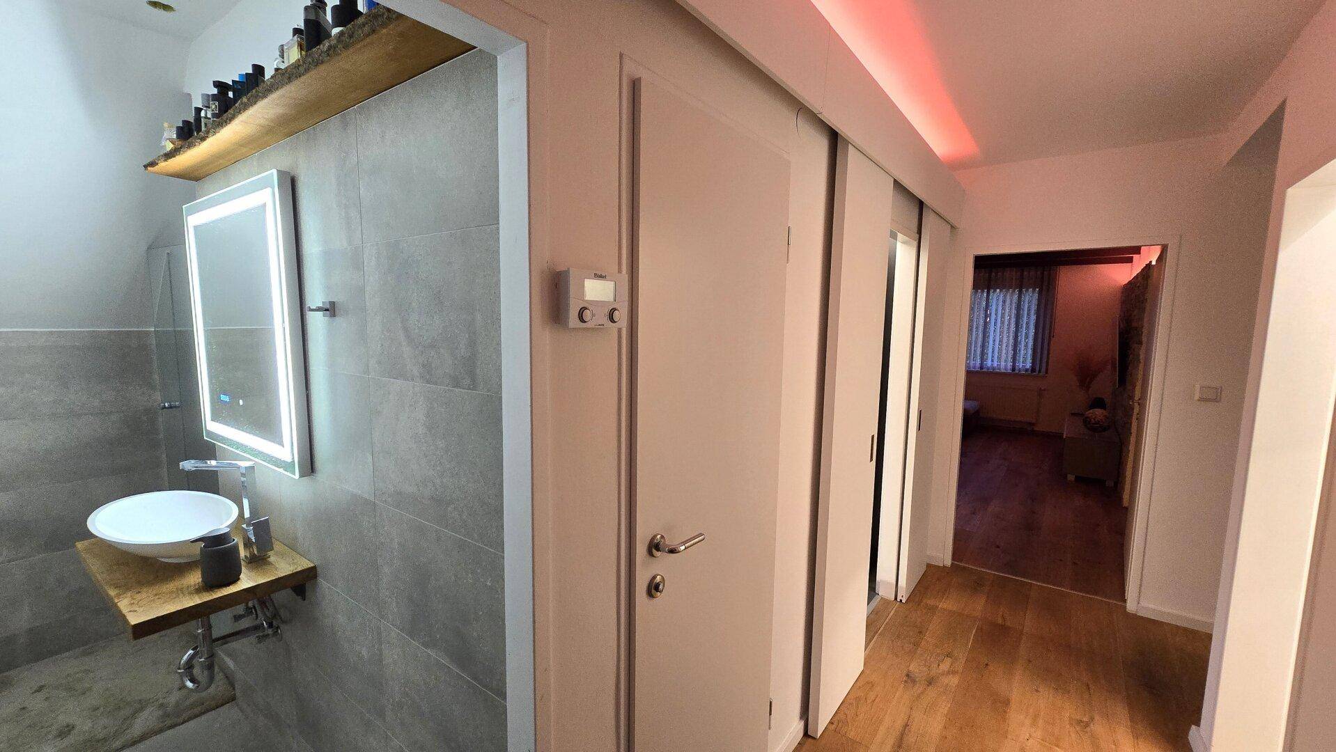 Heller Flur mit Holzboden und Zugang zu einem modernen Badezimmer mit beleuchtetem Spiegel.