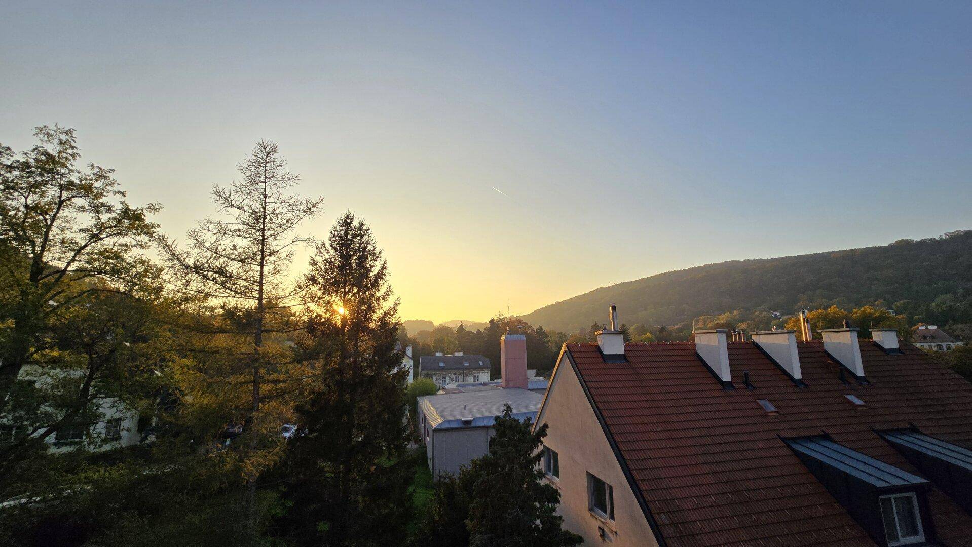 Sonnenuntergang über den Dächern, Bäumen und der hügeligen Landschaft mit warmem Licht.