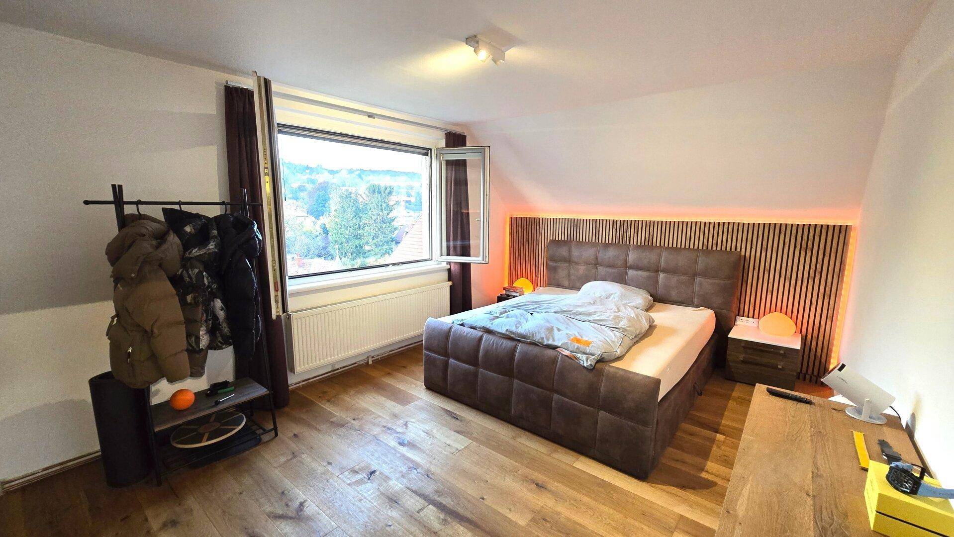 Schlafzimmer im Dachgeschoss mit Holzboden, großem Fenster und indirekter Beleuchtung am Bett.
