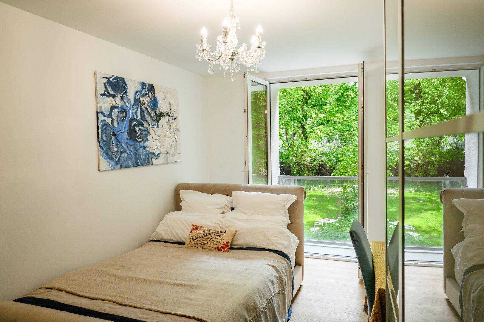 Helles Schlafzimmer mit Einzelbett, Kronleuchter und großem Fenster mit Blick ins Grüne.
