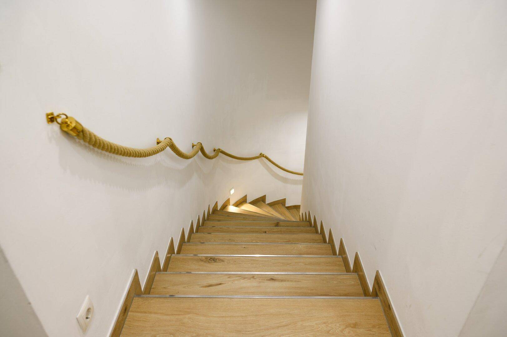 Moderne Holztreppe mit einzigartigem Seilgeländer und weißen Wänden, die nach unten führt.