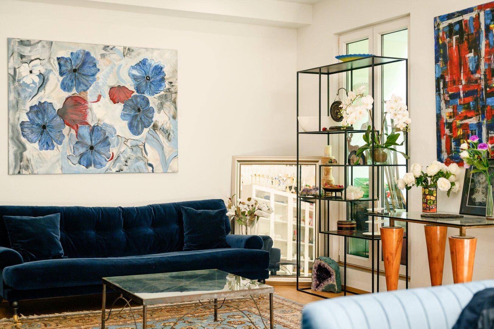 Elegantes Wohnzimmer mit dunkelblauem Samtsofa, Glastisch und farbenfroher moderner Kunst an den Wänden.