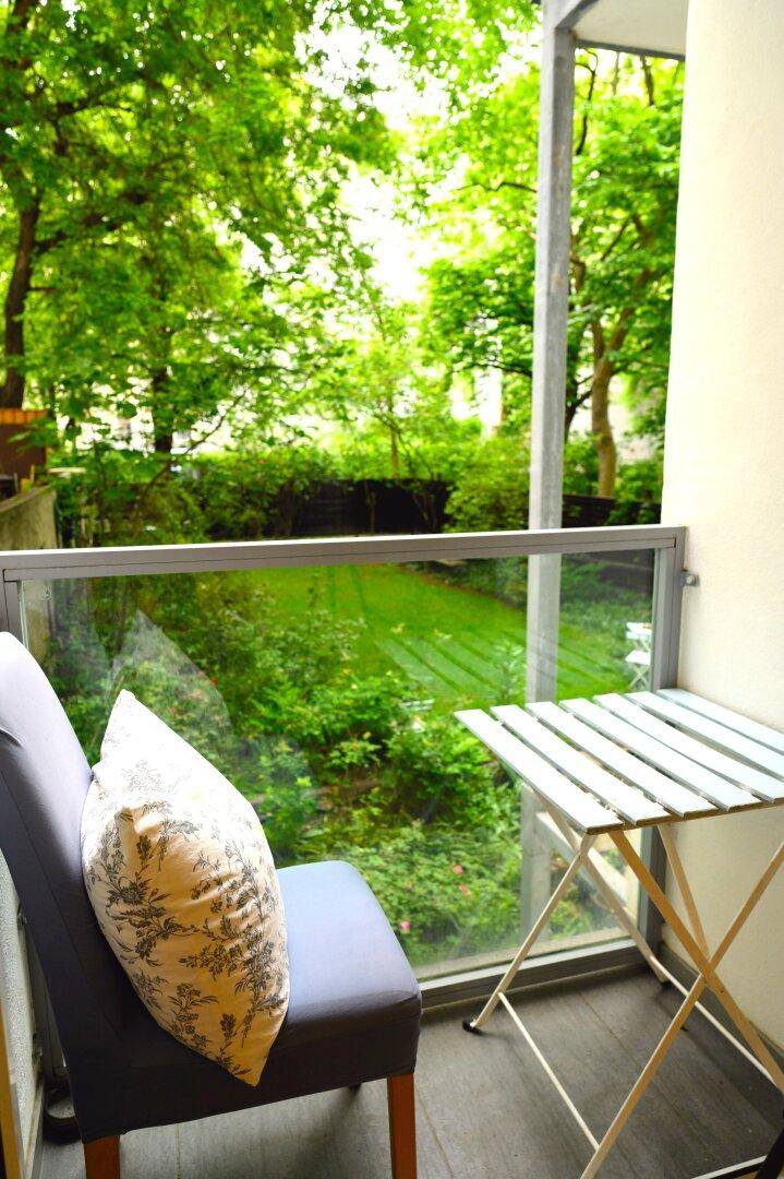 Kleiner Balkon mit Sitzgelegenheit und einem schönen grünen Ausblick auf Bäume und Rasen.