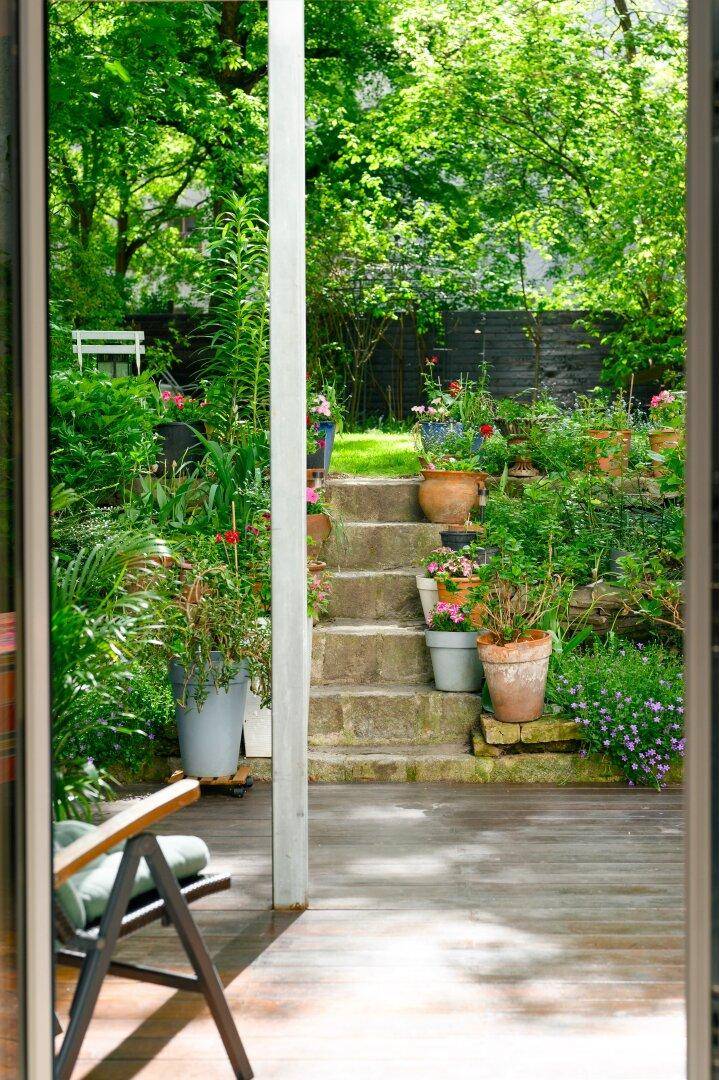 Blick von einer Holzterrasse durch eine Glastür in einen üppigen Garten mit Steinstufen und Topfpflanzen.