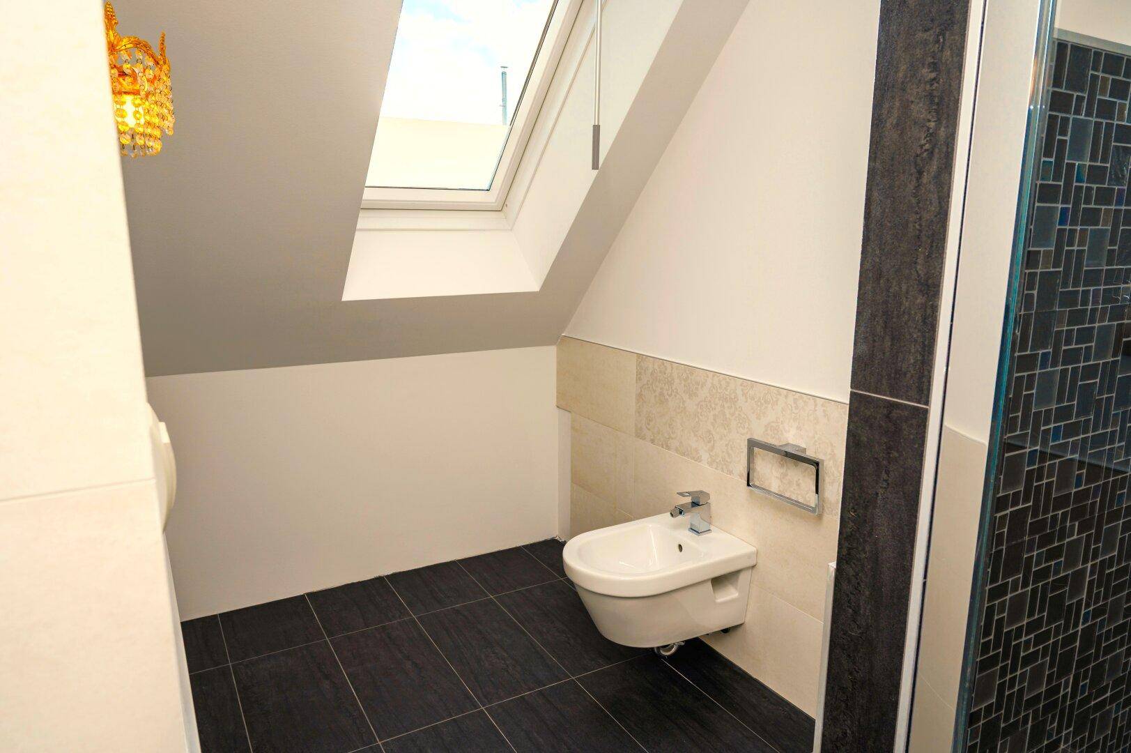 Helles WC mit Bidet, dunklen Bodenfliesen und Dachfenster unter der Schräge.