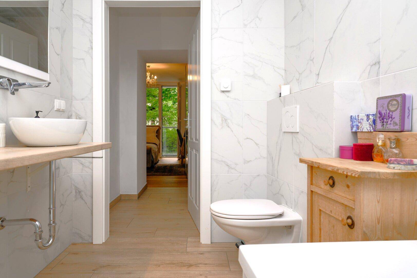 Helles Badezimmer mit Wand-WC, Waschtisch und Marmorfliesen, mit Durchgang zu weiterem Raum.