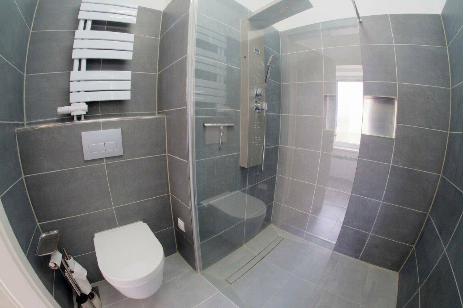 Modernes Badezimmer mit grauen Fliesen, wandhängendem WC und begehbarer Dusche.