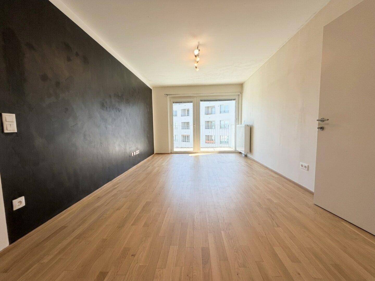 Leeres Wohnzimmer mit dunkler Akzentwand, hellem Parkettboden und Zugang zum Balkon.