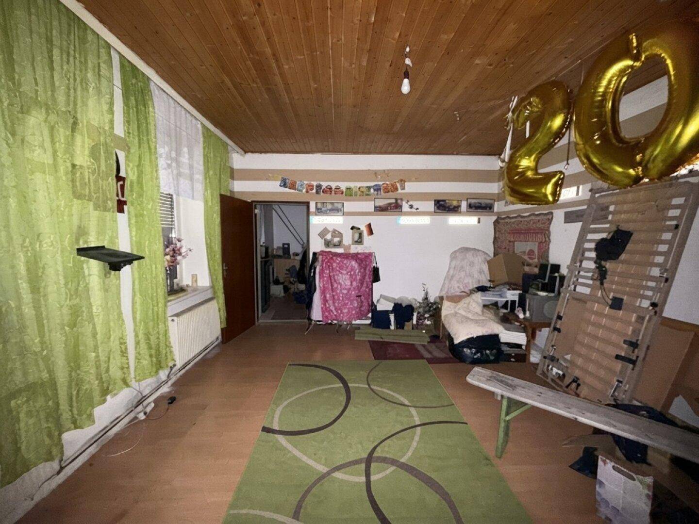 Zimmer mit Holzdecke, grünen Vorhängen und Teppich, derzeit als Abstellraum genutzt.