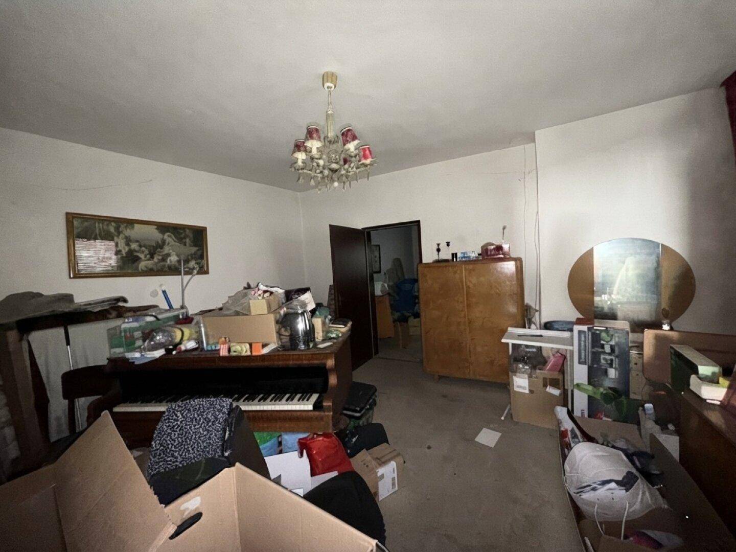 Unaufgeräumtes Wohnzimmer mit Klavier, Möbeln und vielen Kartons, die auf eine Renovierung hindeuten.