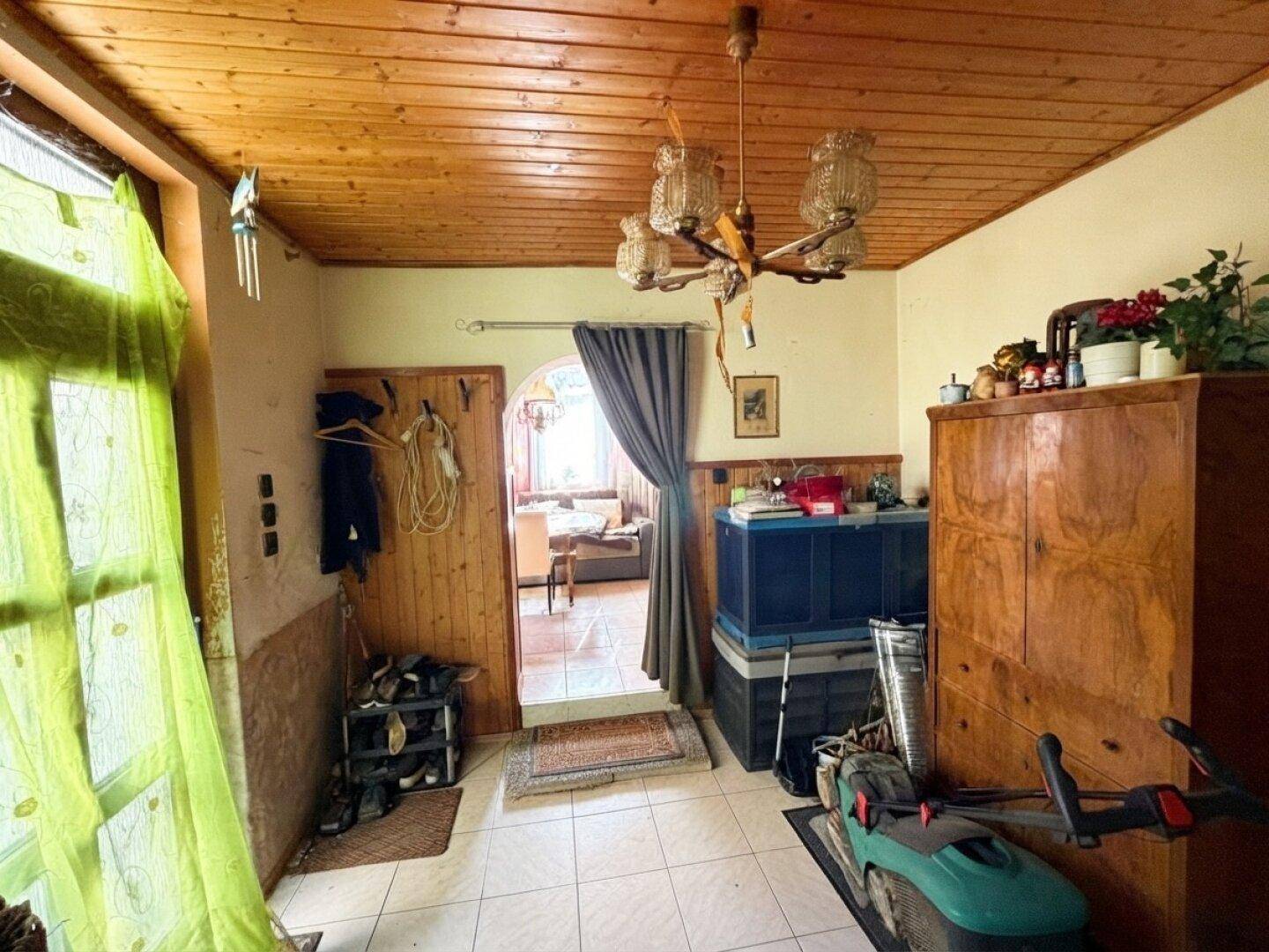 Eingangsbereich mit Holzdecke, Fliesenboden und Blick in einen weiteren Raum, derzeit als Abstellfläche genutzt.