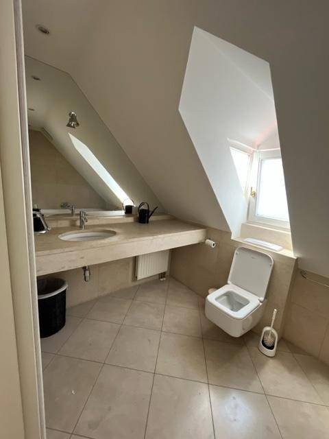 Helles Badezimmer mit beigen Fliesen, wandhängendem WC, Waschtisch und einem Fenster in der Dachschräge.